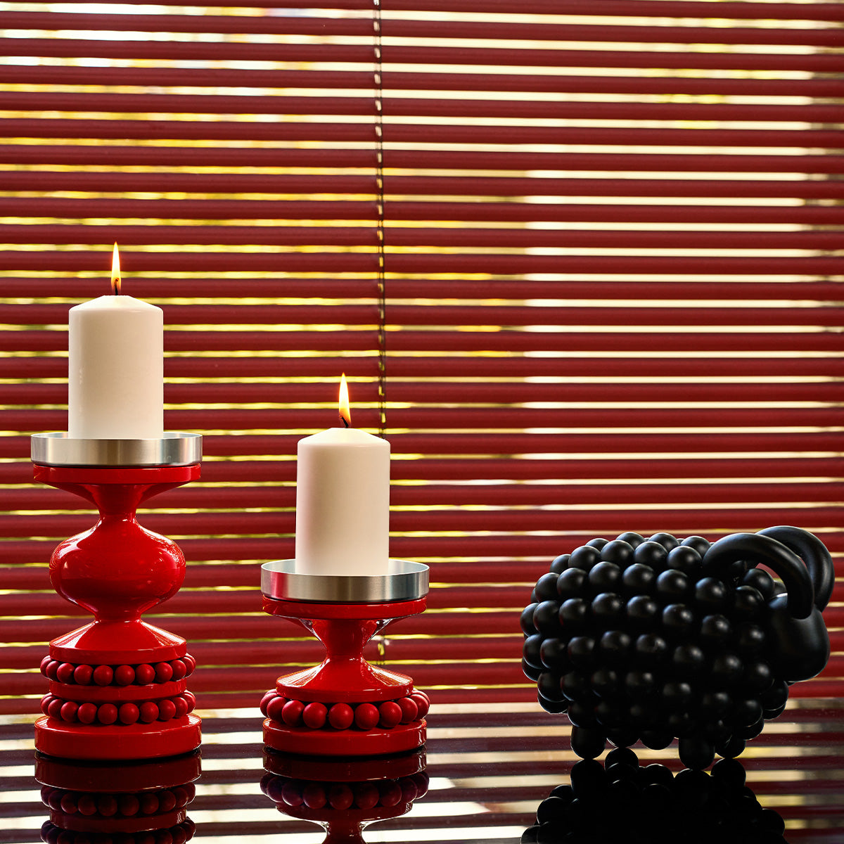 Two red candlestick holders with lit white candles in front of red window blinds, with a black ram figurine on the right.