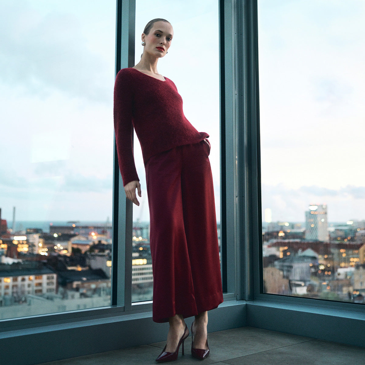 A woman stands by large floor-to-ceiling windows wearing a matching burgundy knit long-sleeve top and skirt set, with burgundy high heels, against a city skyline at dusk.