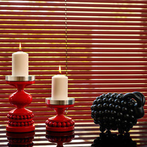 Two red candlestick holders with lit white candles in front of red window blinds, with a black ram figurine on the right.