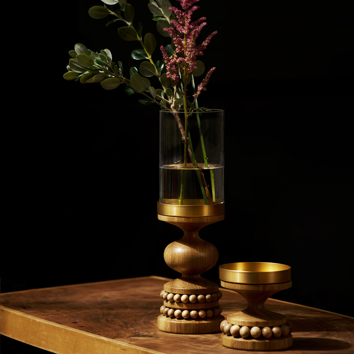 A decorative wooden candle holder with a clear glass cylinder vase and water, a floral arrangement on a wooden surface against a black background.
