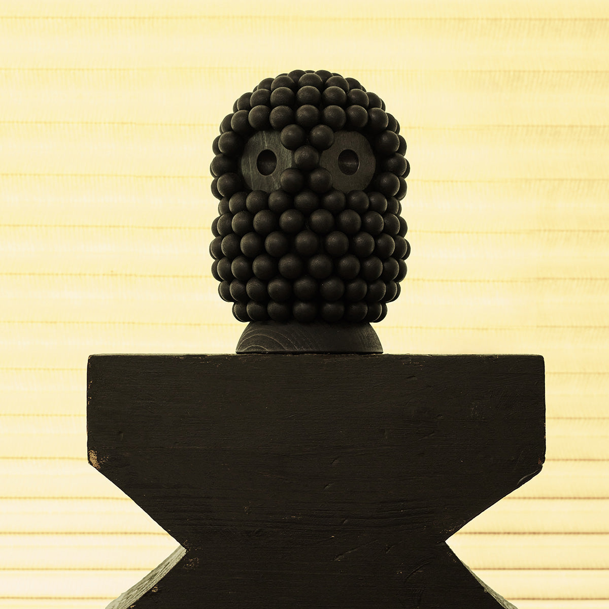 A black decorative sculpture of a stylized owl made of wooden beads, mounted on a dark pedestal against a warm cream-colored paneled background.