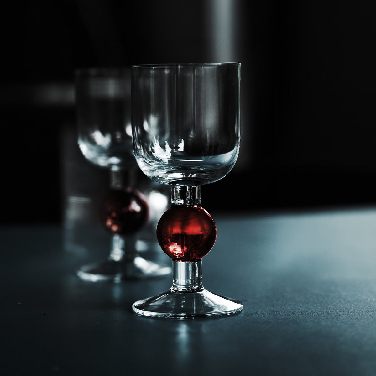 A clear glass goblet with a decorative dark red glass bulb at the base, shown on a dark reflective surface with a second matching goblet blurred in the background.