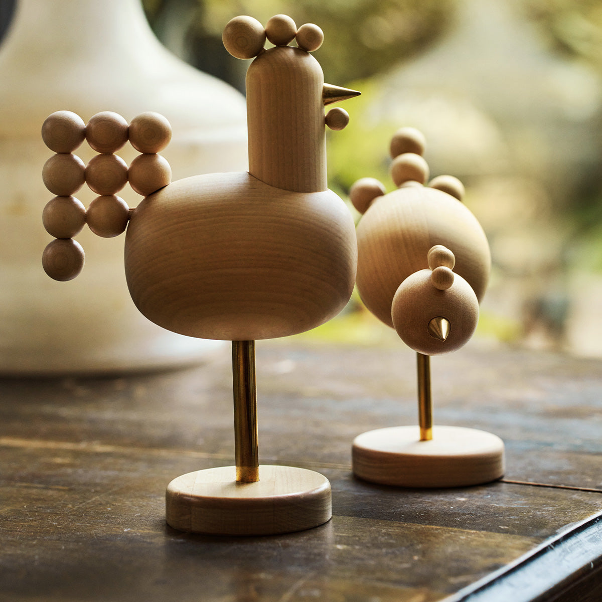 Two minimalist wooden bird figurines on brass stands, displayed on a dark wooden surface with a softly blurred outdoor background.