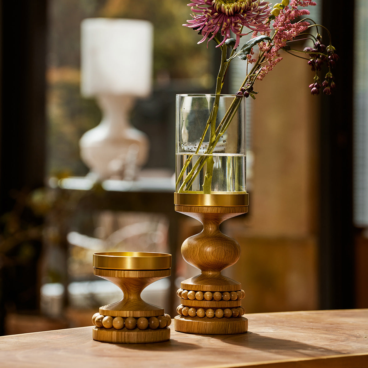 Two decorative wooden candle holders with brass-toned metal tops sit on a wooden table in a softly lit interior. One holder is empty, and the other holds a clear cylindrical glass vase with pink and purple flowers.