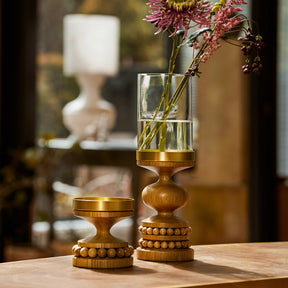 Two decorative wooden candle holders with brass-toned metal tops sit on a wooden table in a softly lit interior. One holder is empty, and the other holds a clear cylindrical glass vase with pink and purple flowers.