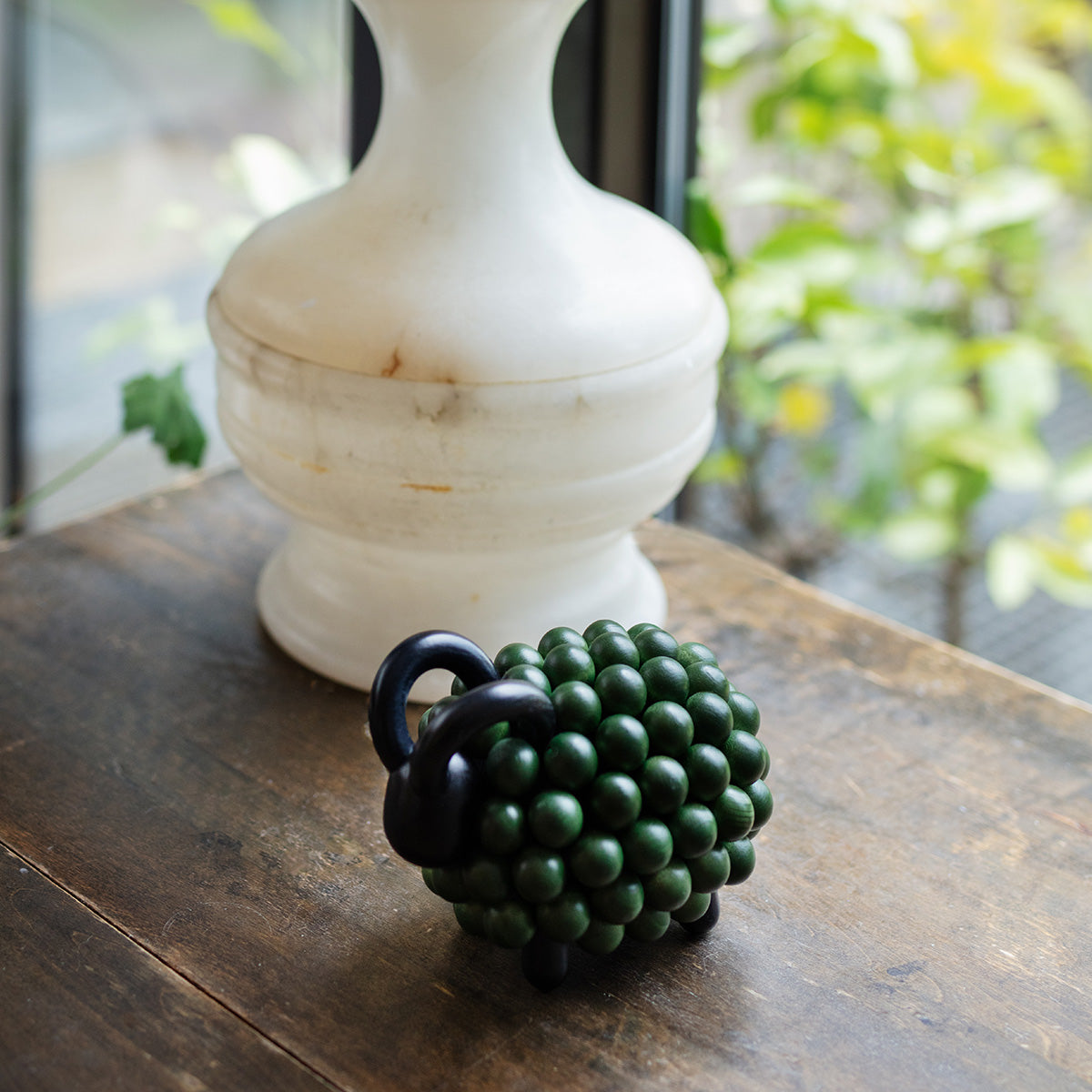 A small green ram figurine sits on a rustic wooden table in front of a white ceramic vase, with a blurred background of green foliage.
